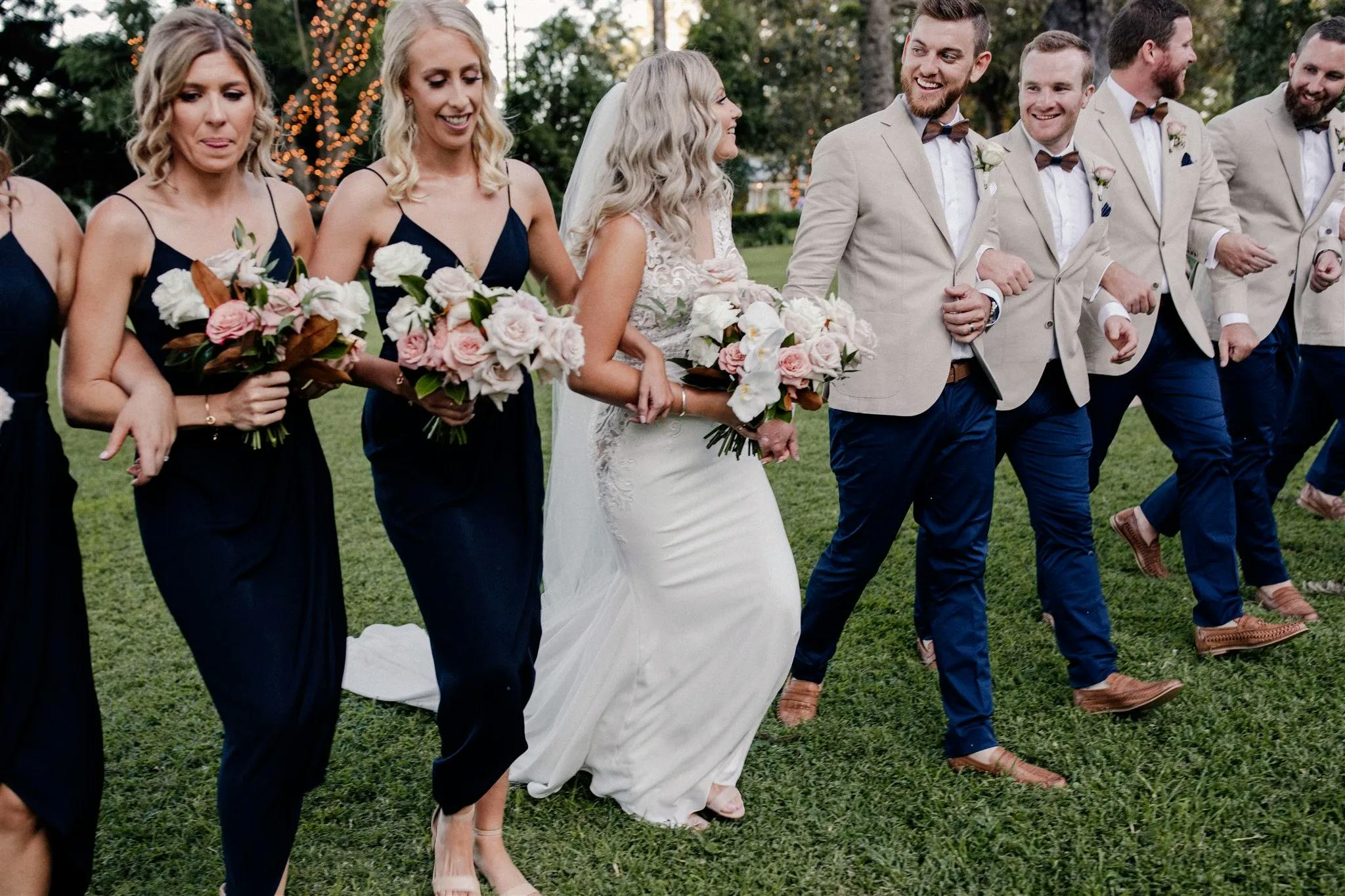 A bride and her bridesmaids, wearing navy blue dresses and holding bouquets of pink and white flowers, walk hand in hand with the groom and his groomsmen, who are dressed in beige jackets, white shirts, bow ties, and blue trousers, on a grassy lawn.