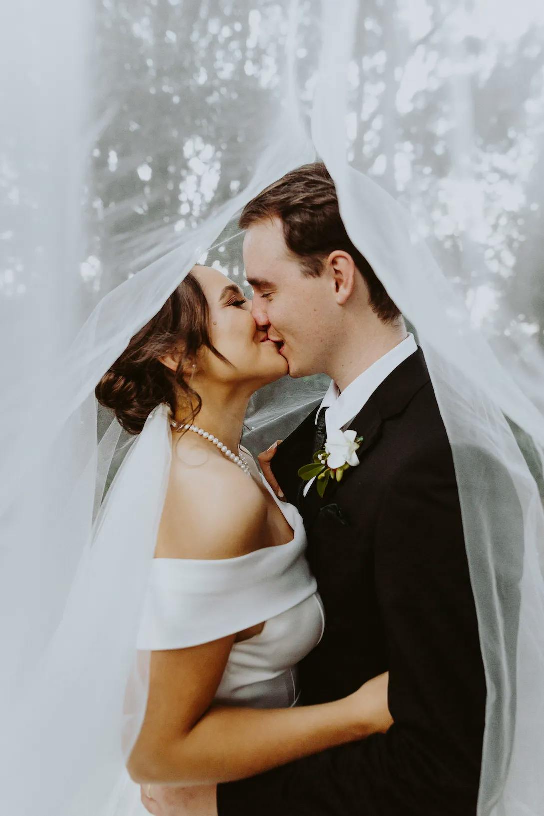 A bride and groom share a kiss under a sheer white veil. The bride is wearing an off-shoulder white gown and pearl necklace, while the groom is in a black suit with a white boutonniere. The soft lighting creates a dreamy, intimate atmosphere.