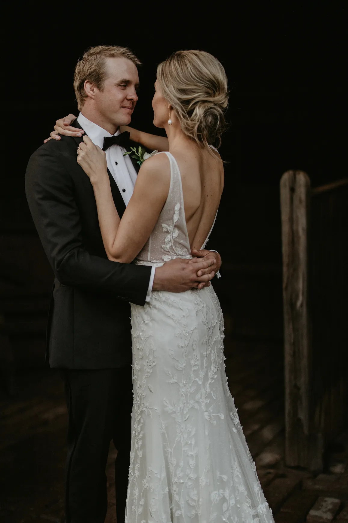 A couple dressed in wedding attire shares an intimate moment. The groom is in a black tuxedo with a bow tie, and the bride wears a white lace dress with a low back. They are gazing into each other's eyes against a dark background. The bride holds the groom's shoulders.