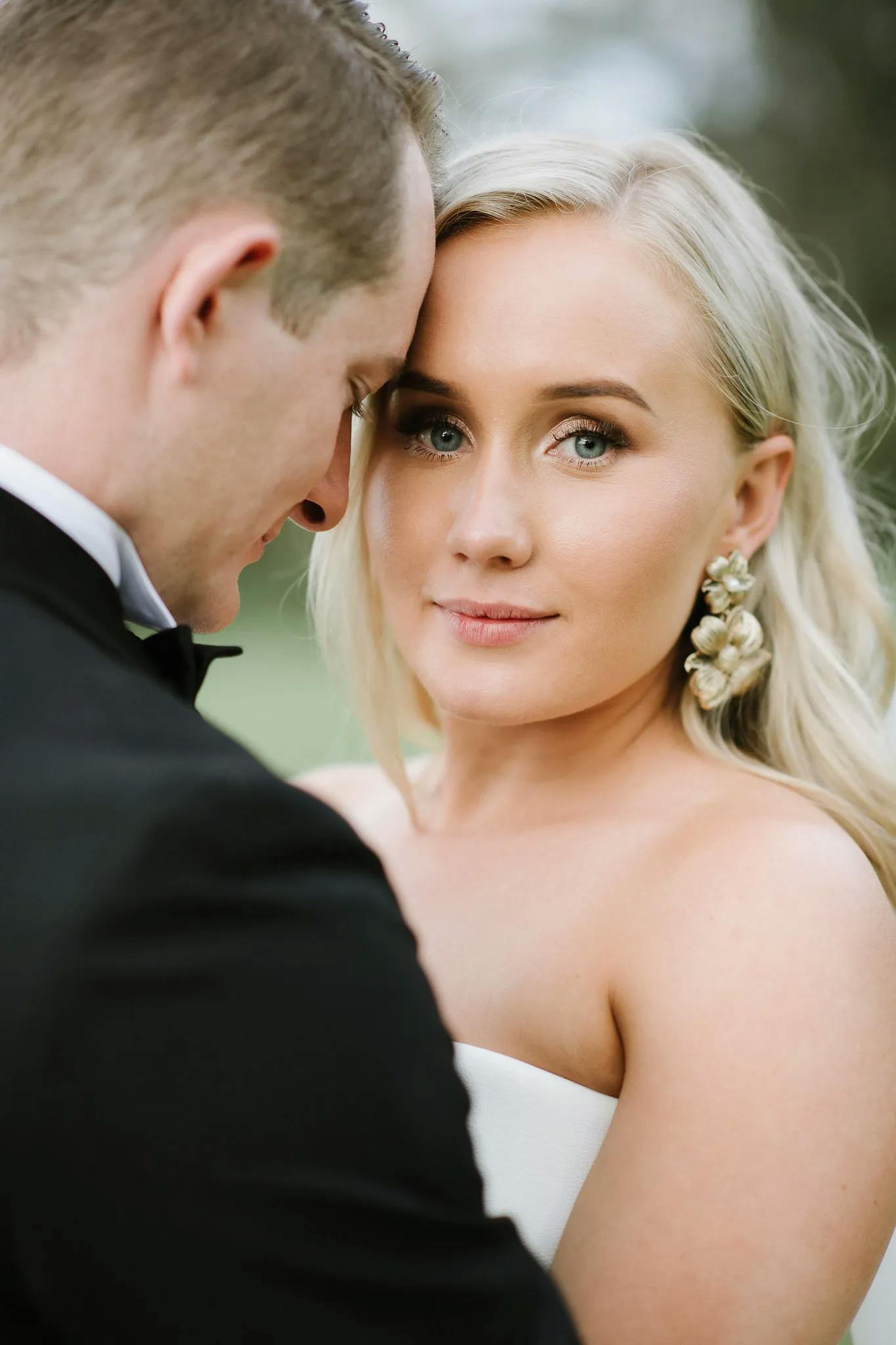 A blonde woman in a strapless white dress looks directly at the camera with a soft smile, while a man in a black suit gently rests his forehead against hers. The background is slightly blurred, emphasizing the couple's intimate moment.