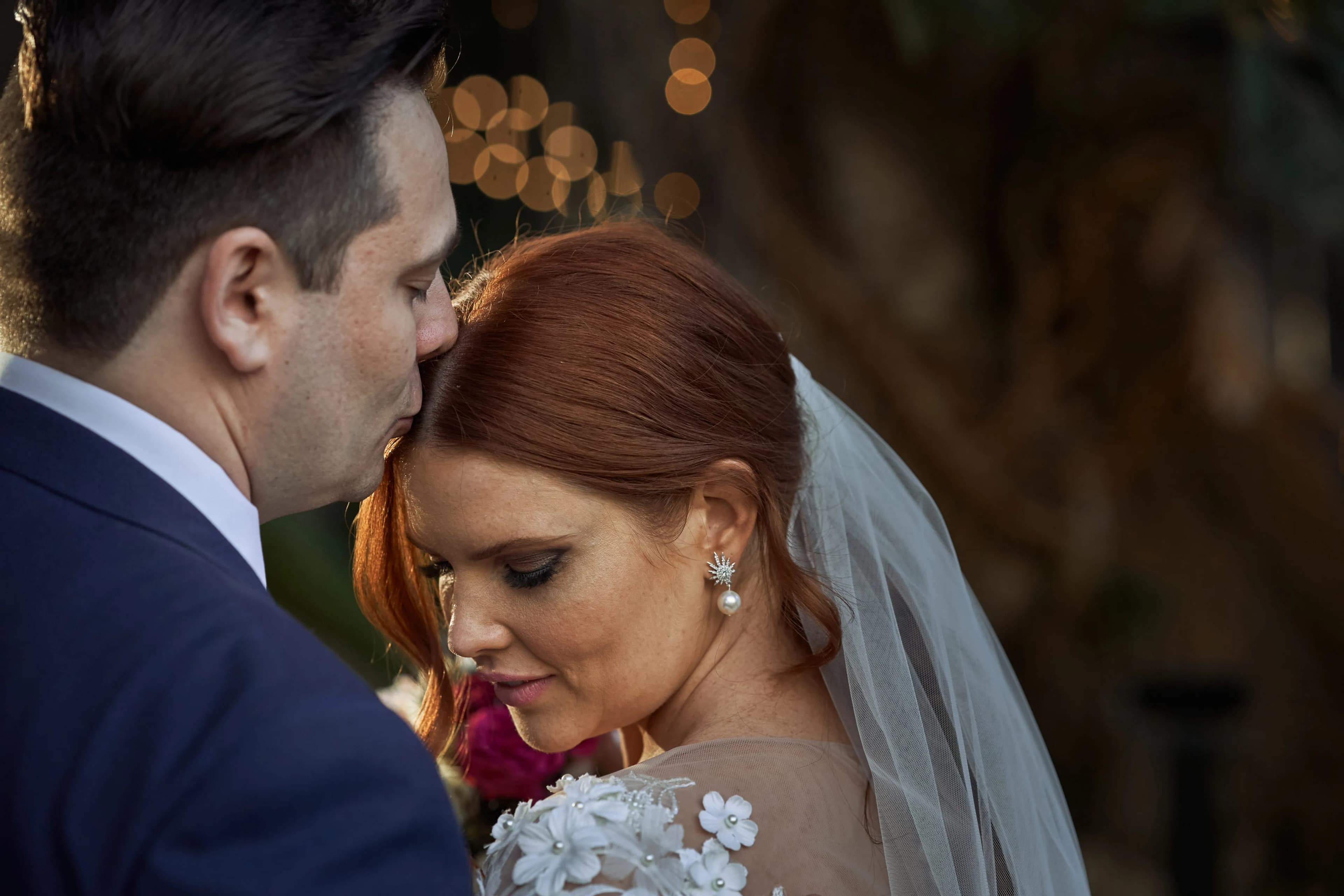 A groom in a navy suit kisses the forehead of his bride, who has red hair and wears a white wedding dress with floral lace details and a veil. She holds a bouquet, and string lights create a warm, romantic background. Both look serene and happy.