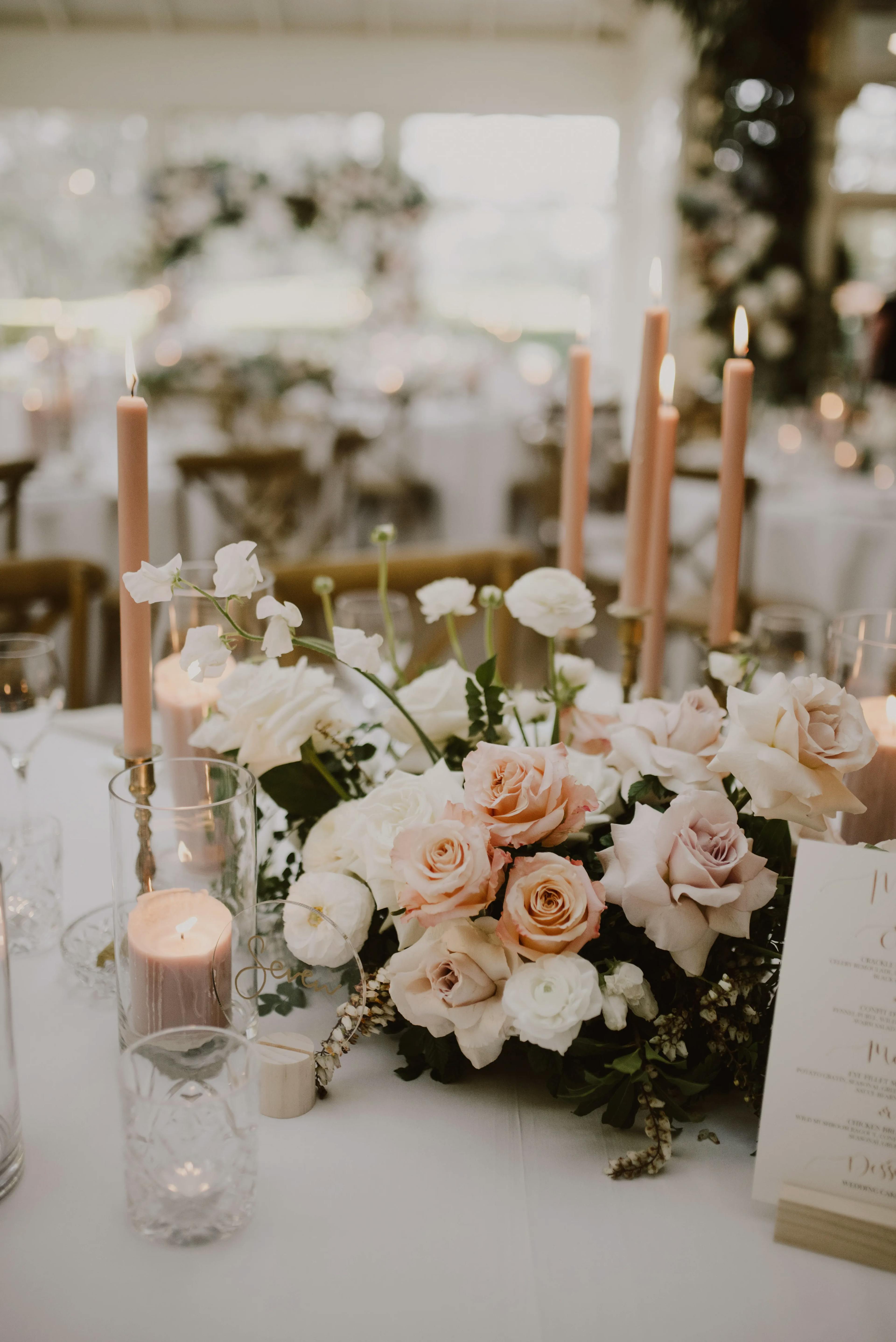 A beautifully arranged table centerpiece features light pink and white roses, lush greenery, and tall, lit pink taper candles in glass holders, creating an elegant and romantic ambiance at an event. The table is set with more roses and candles, enhancing the decor.