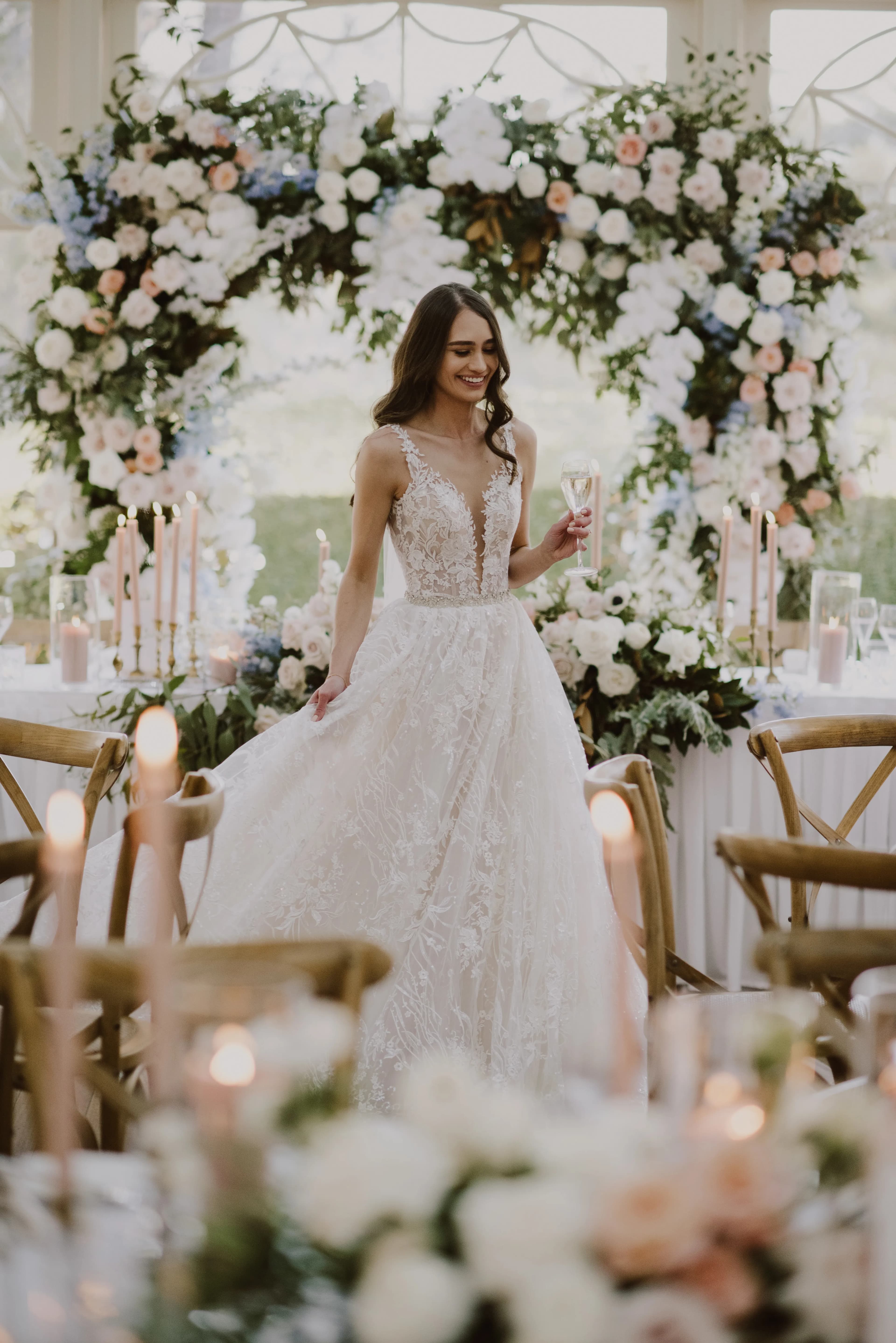 A bride in a white lace wedding gown holds a champagne flute and smiles. She stands before a floral arch adorned with white and pastel flowers with a table set for a wedding feast in front. Candles on the table add a warm, romantic glow to the scene.