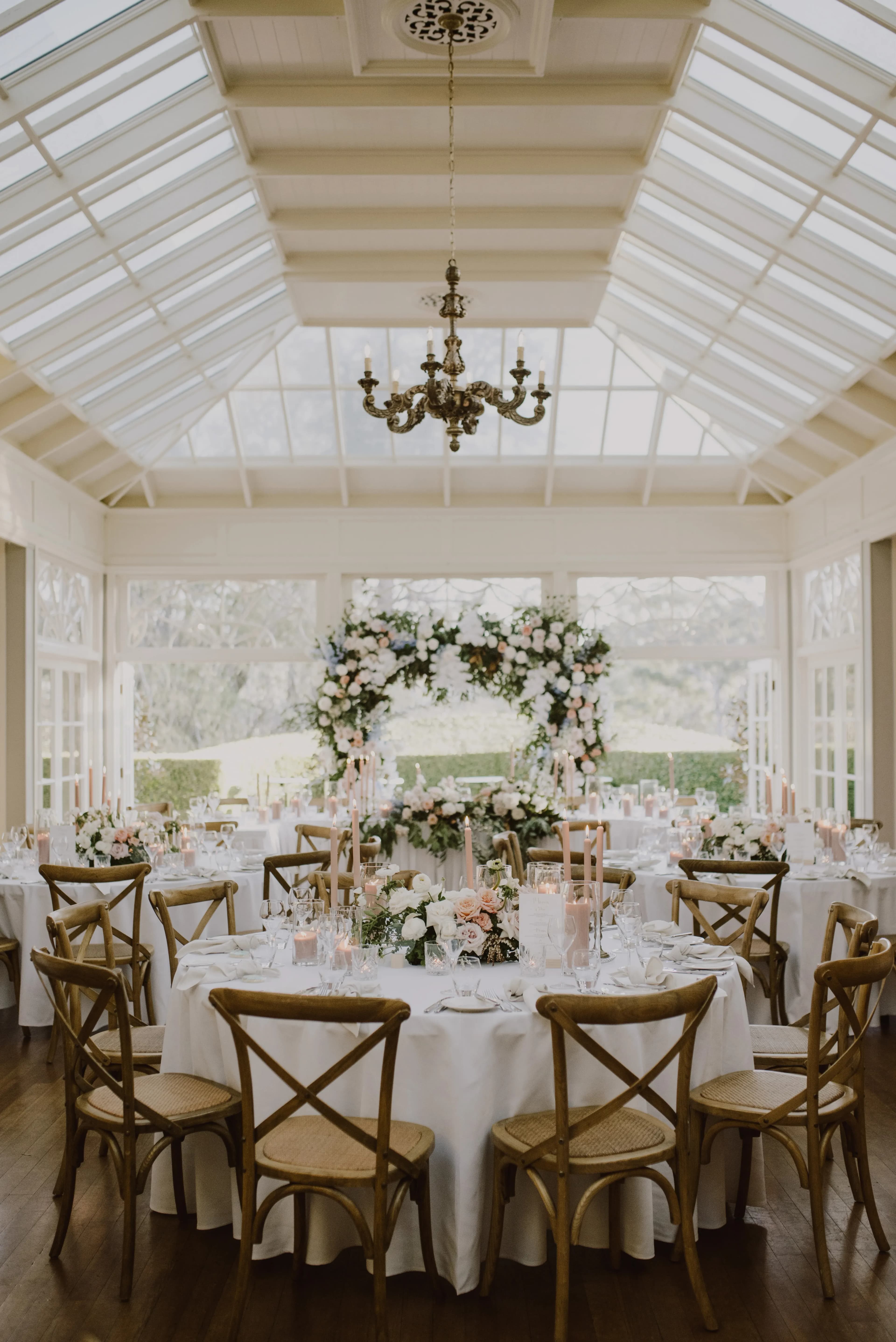 A beautifully decorated wedding reception venue with wooden cross-back chairs and round tables adorned with white tablecloths, floral centerpieces, and candles. An elegant floral arch serves as a backdrop, all under a glass-paneled ceiling enhancing natural light.