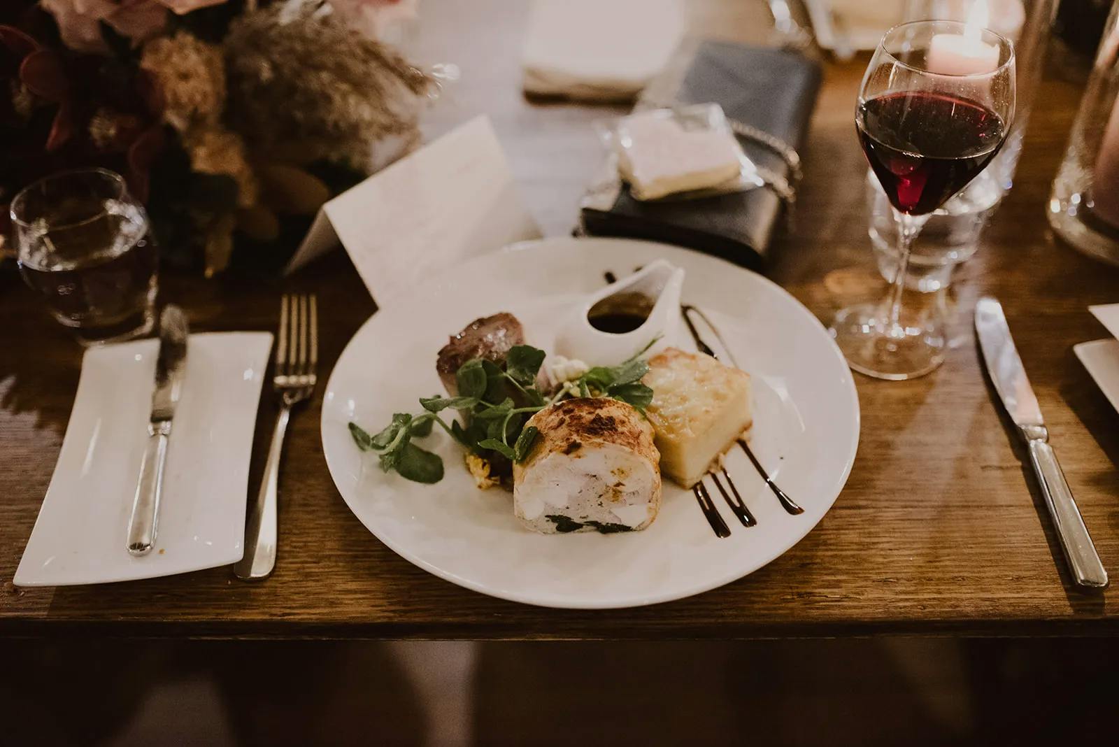 A white plate on a wooden table holds a gourmet meal, including a piece of meat, mashed potatoes, greens, and sauce in a small dish. A glass of red wine, a water glass, cutlery, and a folded napkin are neatly arranged around it. Decor is visible in the background.