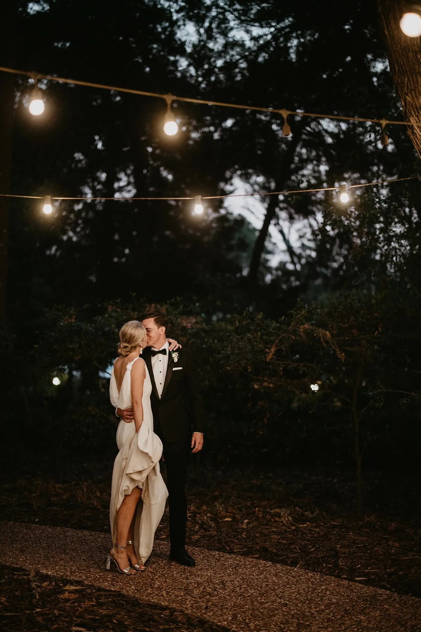 A couple dressed elegantly in wedding attire share a romantic moment under string lights in an outdoor nighttime setting. The woman in a white dress and the man in a black suit stand close, seemingly about to kiss, surrounded by trees and soft lighting.