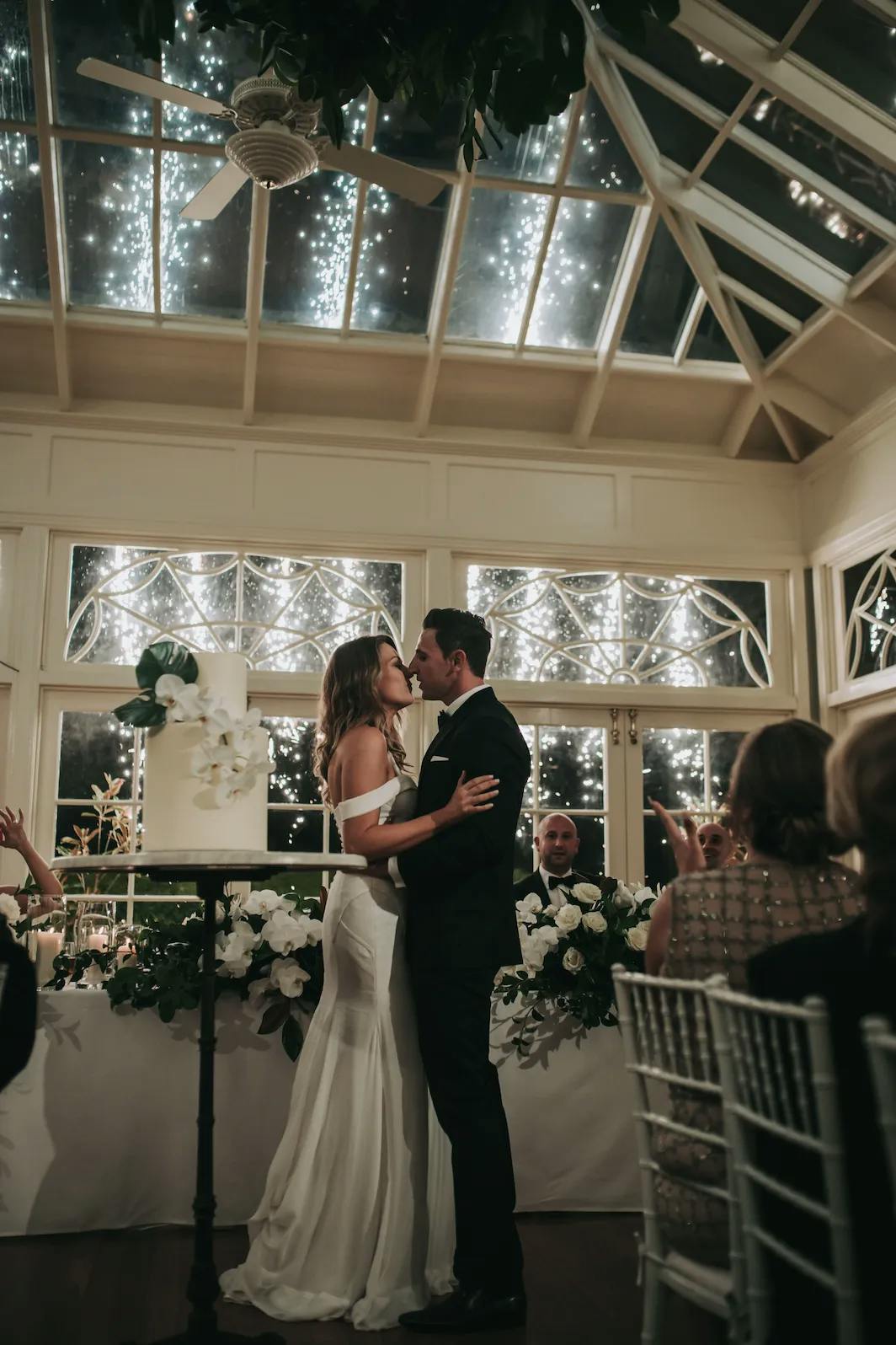 A bride and groom share a kiss in a romantic setting with fireworks lighting up the night sky through a large glass window behind them. They are surrounded by white floral arrangements and seated guests inside a beautifully decorated venue.