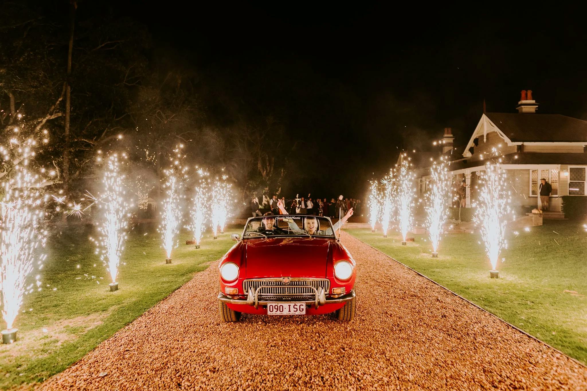 A vintage red convertible drives down a gravel driveway lined with bright sparklers, illuminating the night. The car carries passengers raising their arms in celebration. In the background, people watch from a lawn and a lit house is visible.