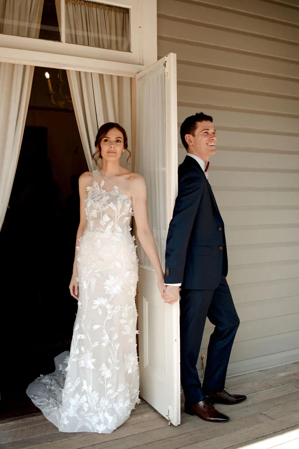 A bride in an elegant white dress and a groom in a dark suit stand back-to-back, holding hands, in front of a house with light-colored wooden siding. Both look in different directions, the groom smiling and the bride with a serene expression.