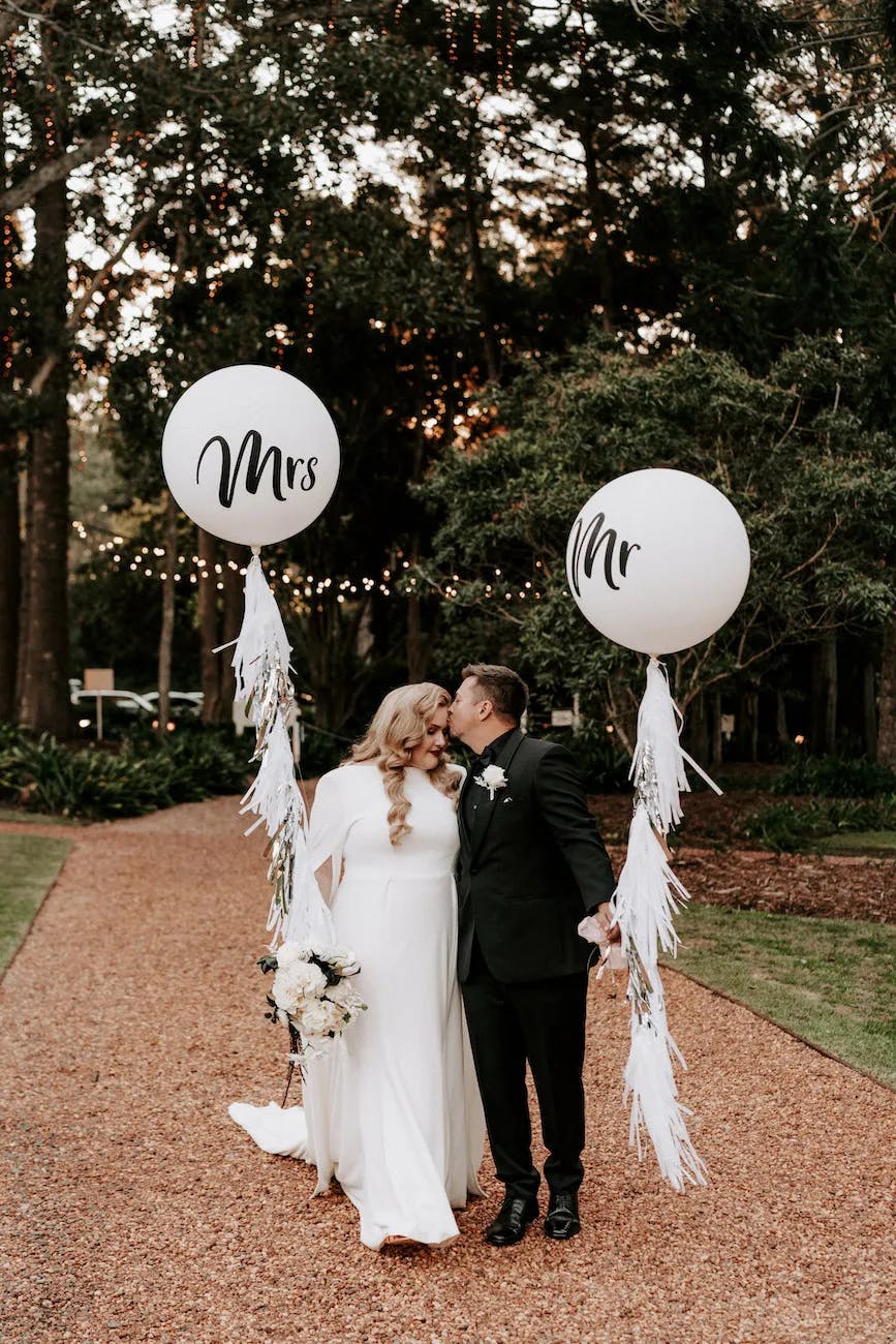 A newlywed couple shares a kiss while holding large white balloons that read "Mr" and "Mrs." The bride, in a flowing white gown, holds a bouquet of flowers. They stand on a gravel path surrounded by lush greenery and string lights, creating a romantic setting.