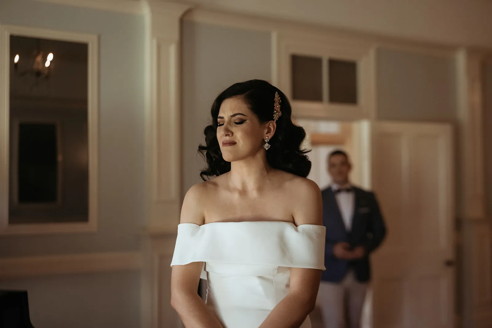A woman in an off-shoulder white dress looks emotional with her eyes closed. She stands in a room with light-colored walls and paneling. A man in a suit stands in the background, slightly out of focus, by a door.
