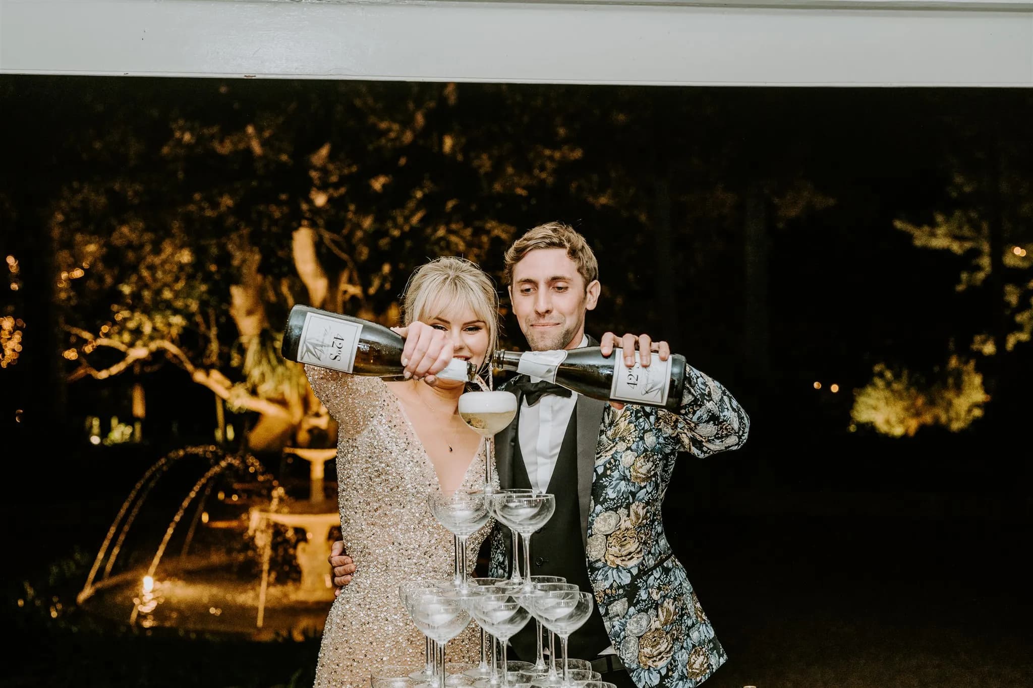 A smiling couple is pouring wine from two bottles into a stack of champagne glasses. The woman is wearing a sparkly dress, and the man is wearing a floral patterned suit jacket. They are standing outdoors at night with a fountain and trees softly lit in the background.