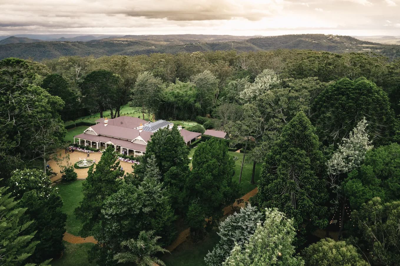 Aerial view of a large, elegant house with a red roof surrounded by dense forest and gardens. A winding path leads to the house, and mountains are visible in the distance under a cloudy sky.