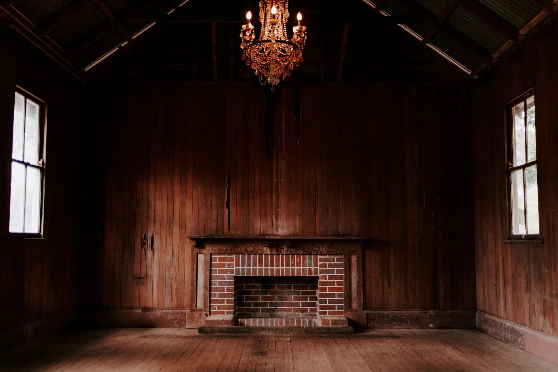 A dimly lit, empty room with wooden walls and flooring. A brick fireplace is centered on the far wall. Above the fireplace hangs an ornate chandelier. Two windows on either side of the room allow minimal light to enter, casting shadows across the space.