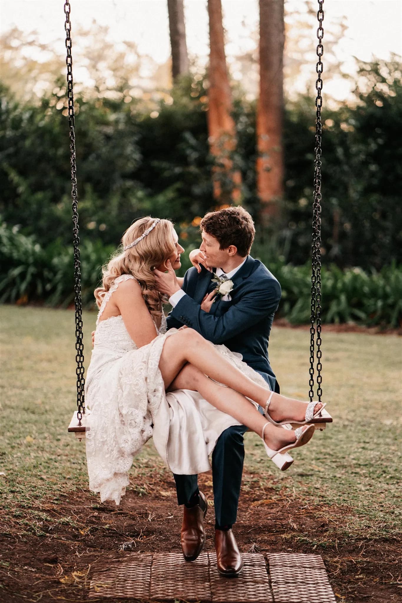 A bride in a white lace dress and a groom in a navy suit sit closely on a wooden swing suspended with chains. They look into each other's eyes lovingly, surrounded by greenery and tall trees in the background. The bride's dress and shoes stand out in the serene outdoor setting.