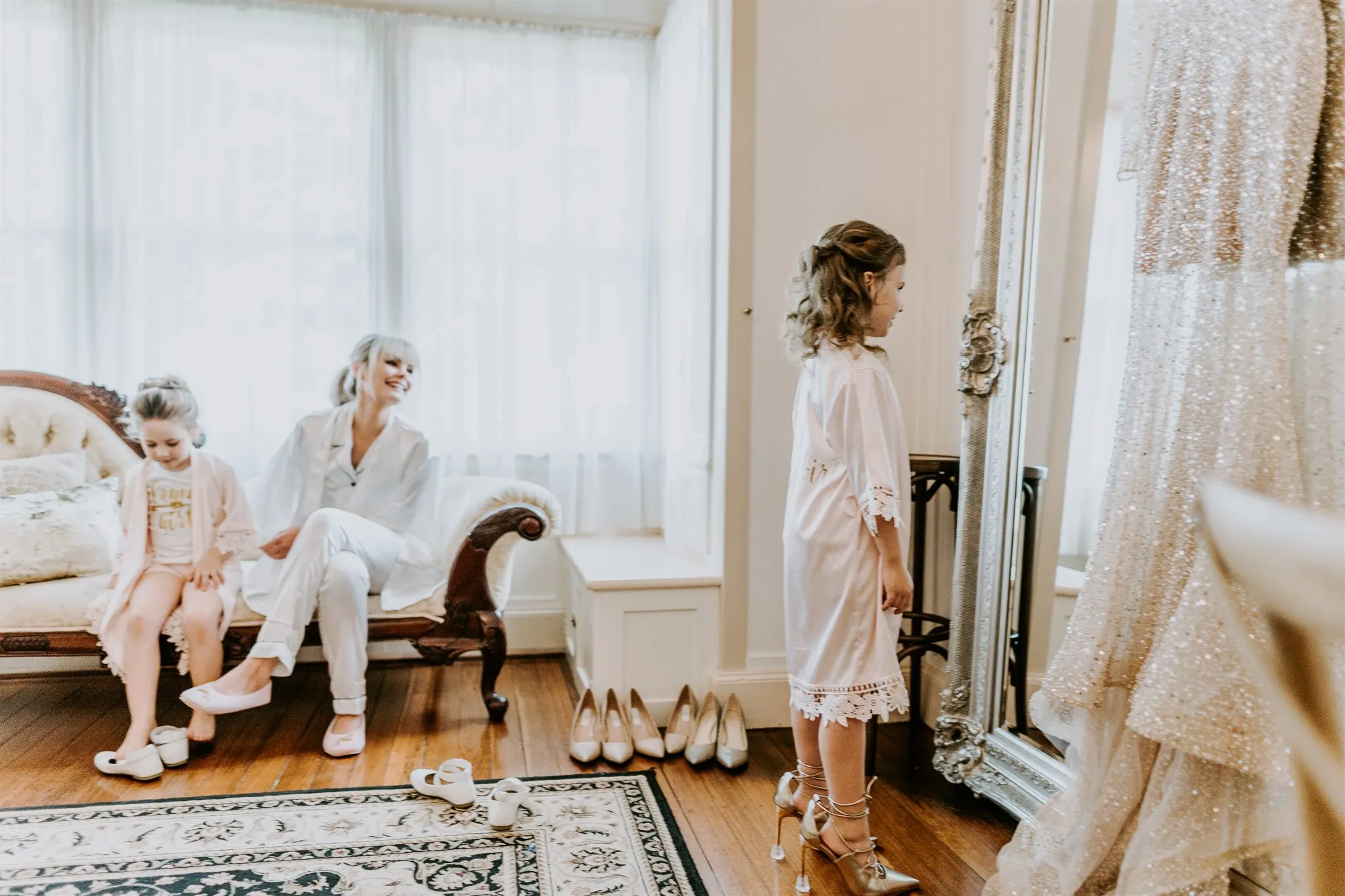 A young girl in a white dress and heels looks at herself in a mirror while an older woman and another young girl sit on a nearby sofa, smiling and watching. Multiple pairs of shoes are lined up on the floor, and the room has a large window with sheer curtains.