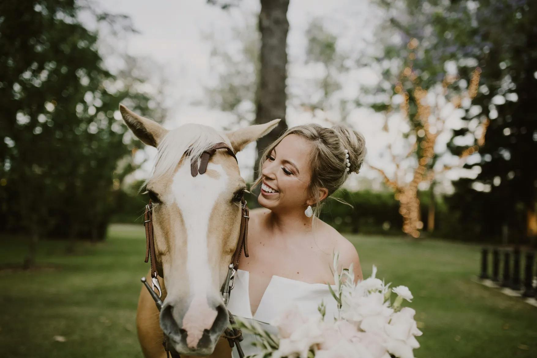 A bride in a white dress is standing outdoors, smiling and holding a bouquet of flowers while affectionately leaning close to a light brown horse. The background features greenery and trees with twinkling lights.