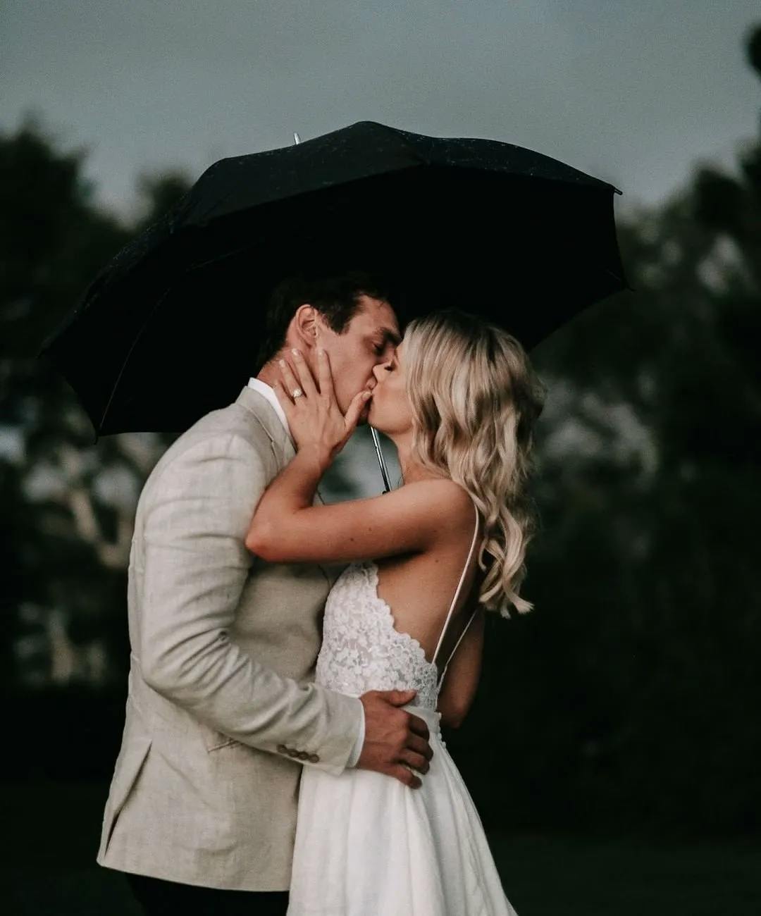 A couple dressed in wedding attire shares a kiss under a black umbrella. The groom, wearing a light-colored suit, gently holds the bride, who is in a white lace dress. The scene is set outdoors with a dark, cloudy sky and blurred greenery in the background.