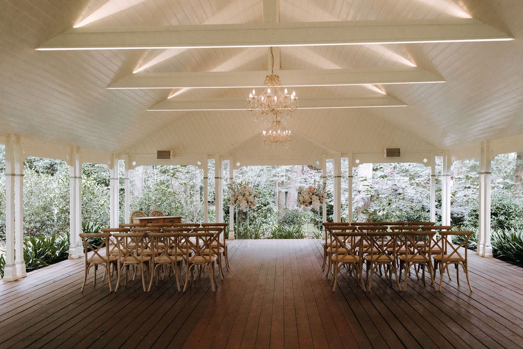 An elegant outdoor wedding ceremony setup under a white pavilion with a wooden floor. Wooden chairs arranged in rows face a central area. A crystal chandelier hangs from the vaulted ceiling. Surrounding lush greenery enhances the serene, romantic ambiance.