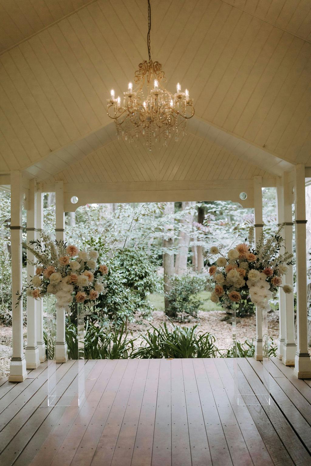 An elegant wedding altar setting featuring a wooden pavilion adorned with a golden chandelier hanging from the ceiling. Two large, flower arrangements with white and pastel flowers stand on transparent pedestals on either side of the altar area, framed by greenery.