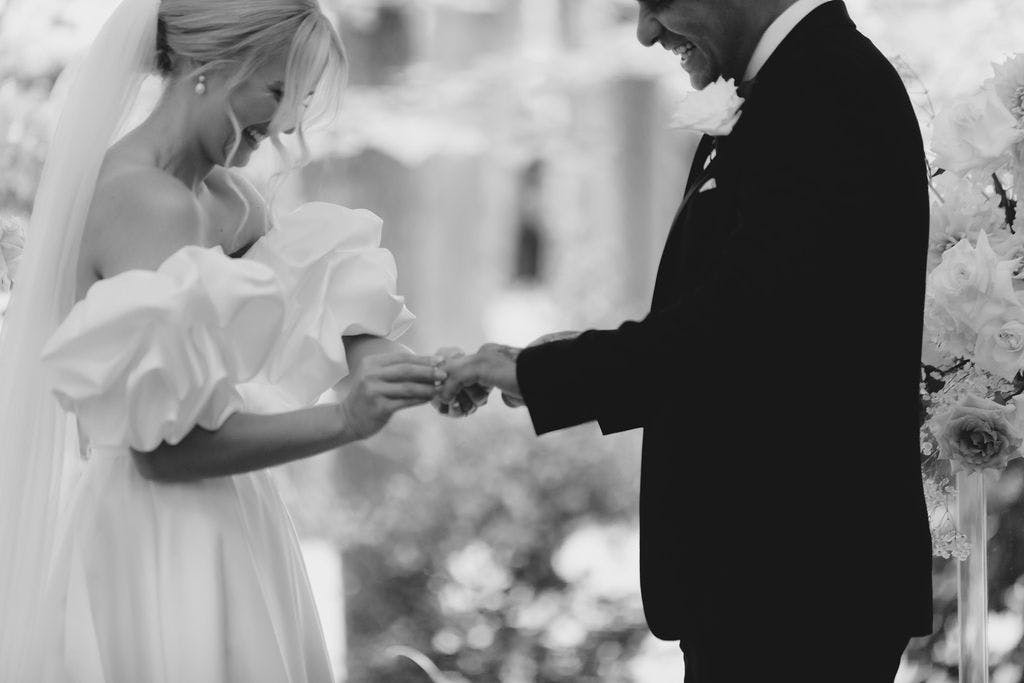 A black and white photo of a wedding ceremony. A bride with a veil and off-the-shoulder dress is placing a ring on the groom's finger. The groom, in a suit with a boutonniere, smiles joyfully. The background features soft, out-of-focus floral decorations.