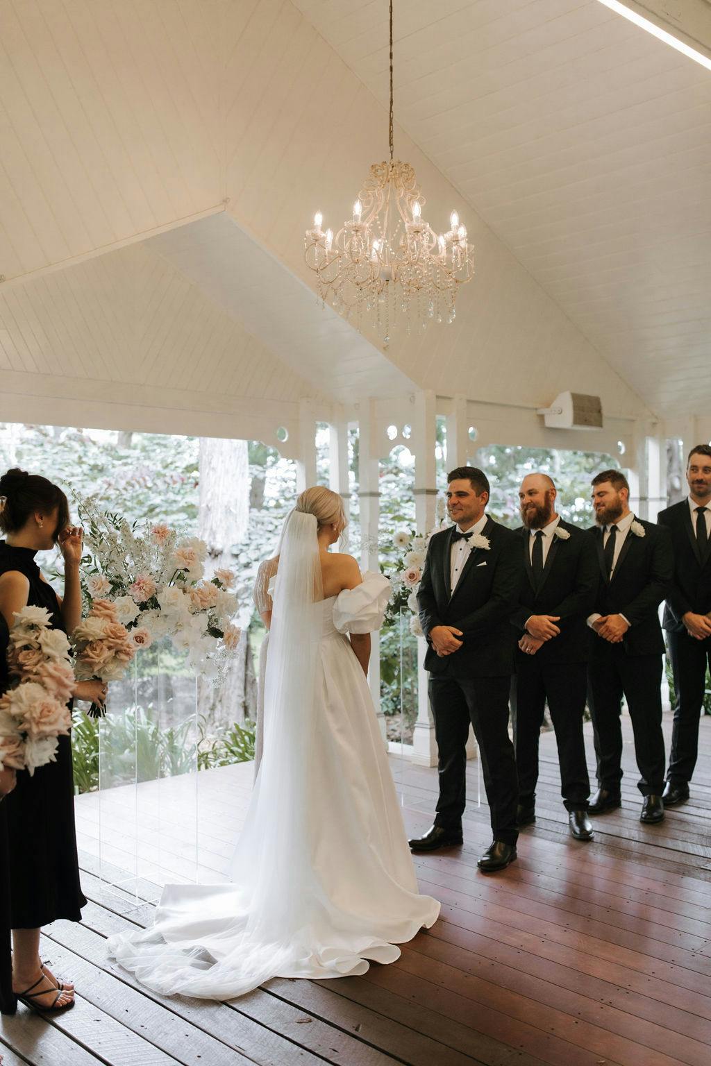 A bride in a white wedding gown stands facing the groom, who is dressed in a black tuxedo, at an indoor ceremony. The groom smiles with groomsmen in black suits standing beside him. A bridesmaid holds flower arrangements on the left, beneath a chandelier.