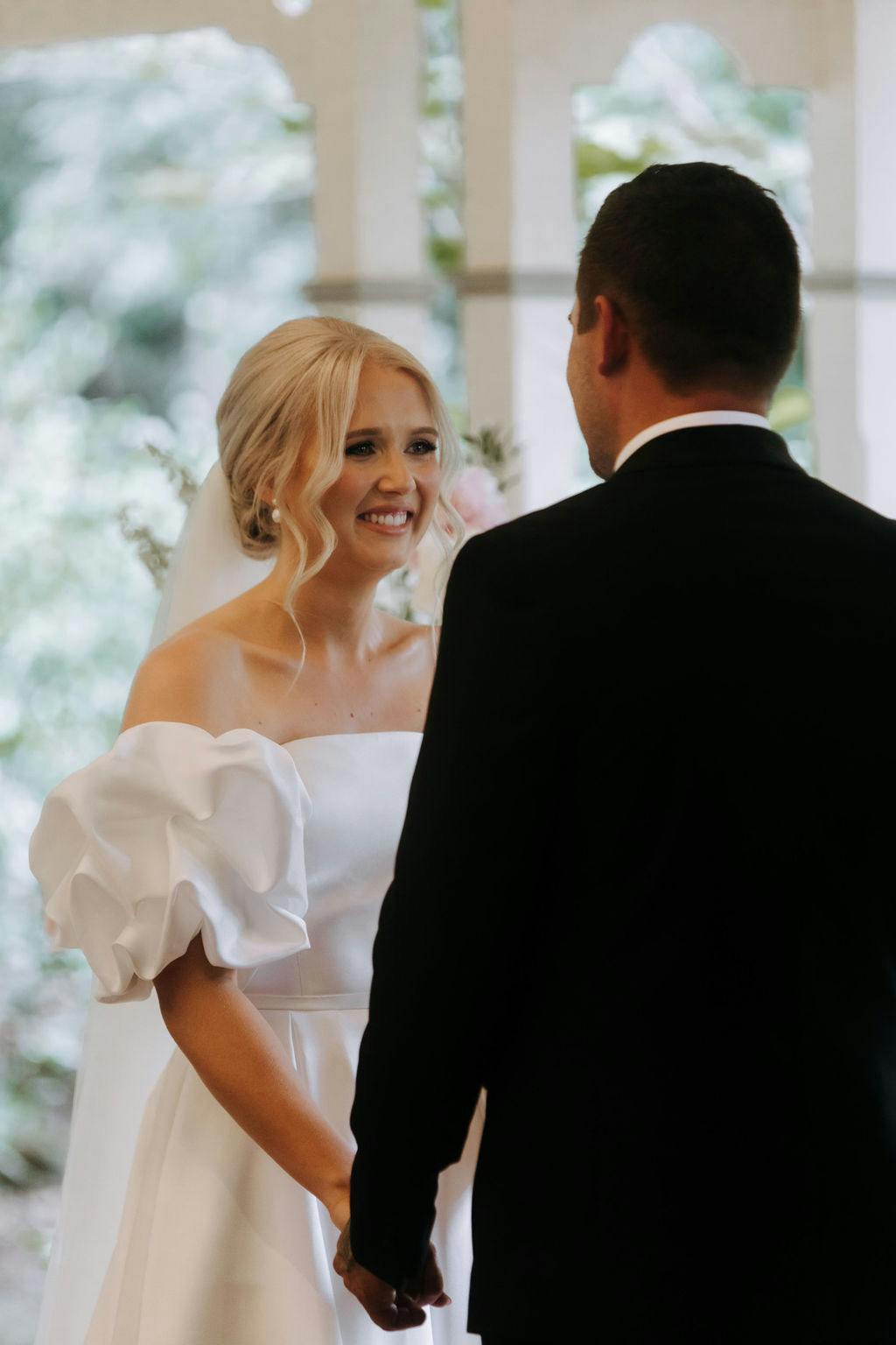 A bride and groom are standing face to face, holding hands. The bride is wearing an off-the-shoulder white wedding dress with puff sleeves, and her hair is styled in loose curls. The groom is in a black suit. They are smiling at each other in an outdoor setting.