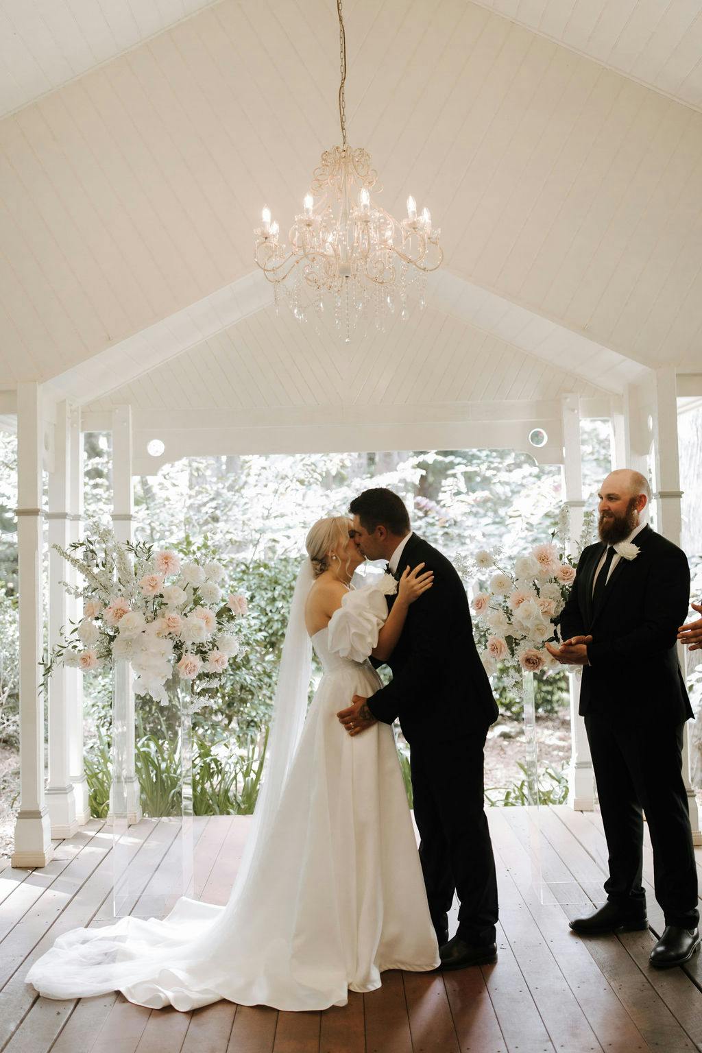 A couple sharing their first kiss at a wedding ceremony under a white pavilion with a chandelier hanging above. They are surrounded by elegant floral arrangements, and a person in a black suit and tie stands to the right, smiling.