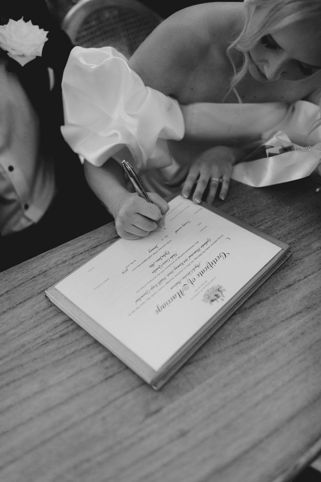 A black and white photo of a bride signing a marriage certificate on a wooden table. She is wearing a wedding dress with puffed sleeves and has blonde hair. A man in a suit, partially visible, is seated next to her.