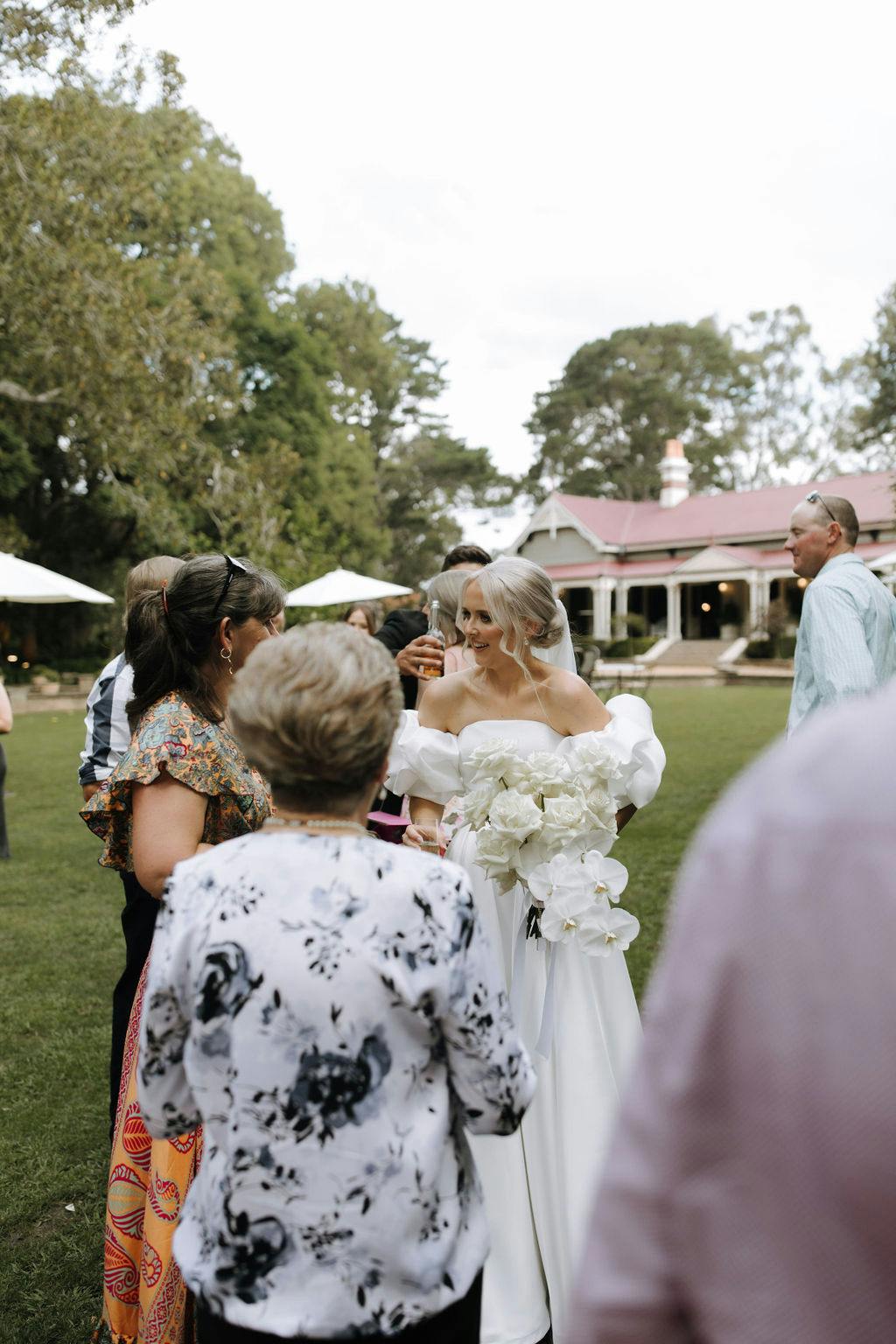 A bride in a white off-the-shoulder gown, holding a bouquet of white flowers, stands outdoors surrounded by guests. Green lawns and trees are visible, along with a building with a red roof in the background. The setting appears to be a garden or park.