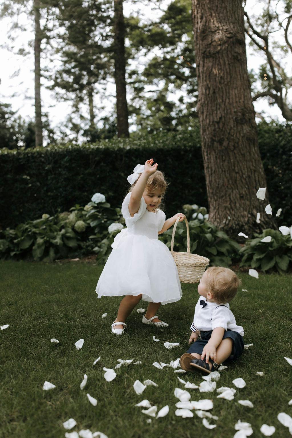 A young girl in a white dress tosses flower petals from a basket while a toddler in a white shirt and dark shorts sits on the grass, surrounded by more petals. They are in an outdoor garden area with trees and greenery in the background.