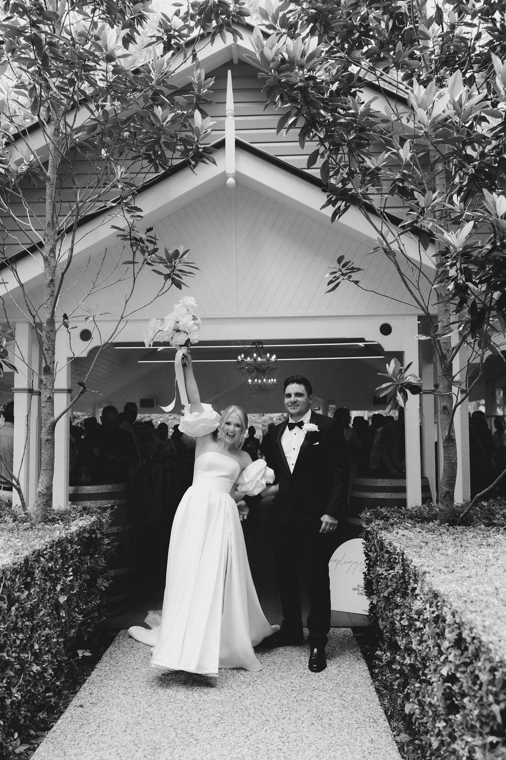 A bride in a white gown and a groom in a tuxedo stand joyfully outside a small chapel adorned with greenery. The bride holds a bouquet high in celebration while the groom smiles beside her. Trees frame the pathway leading to the chapel.