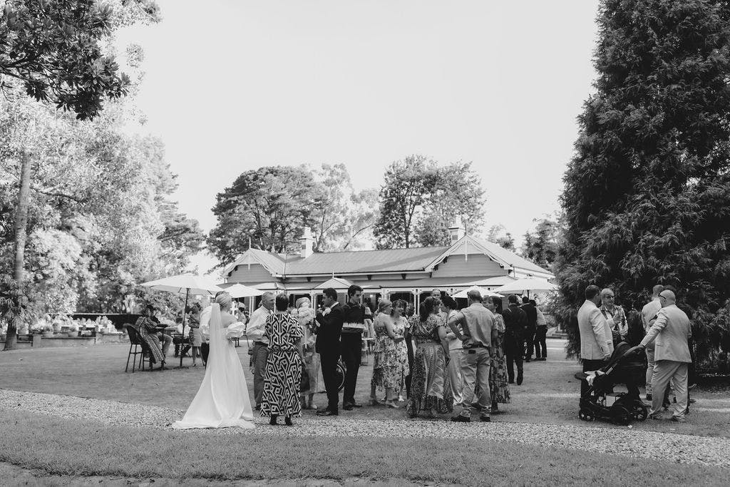 A black-and-white photo of an outdoor wedding reception. Guests are gathered on a grassy area near a large house, socializing and dressed in formal attire. Trees and shrubs surround the space, and a bride in a long gown is seen on the left side of the image.