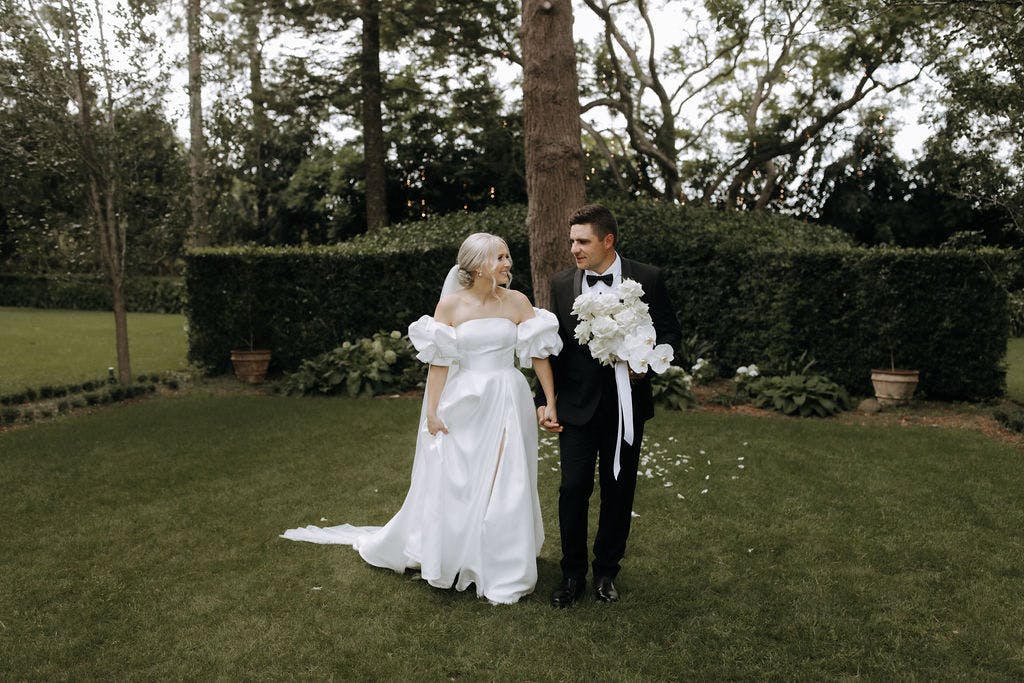 A couple in formal wedding attire walk hand in hand in a lush garden. The bride wears an off-the-shoulder white dress and the groom, a black tuxedo. The groom holds a bouquet of white flowers. They both look happy and the greenery surrounds them.