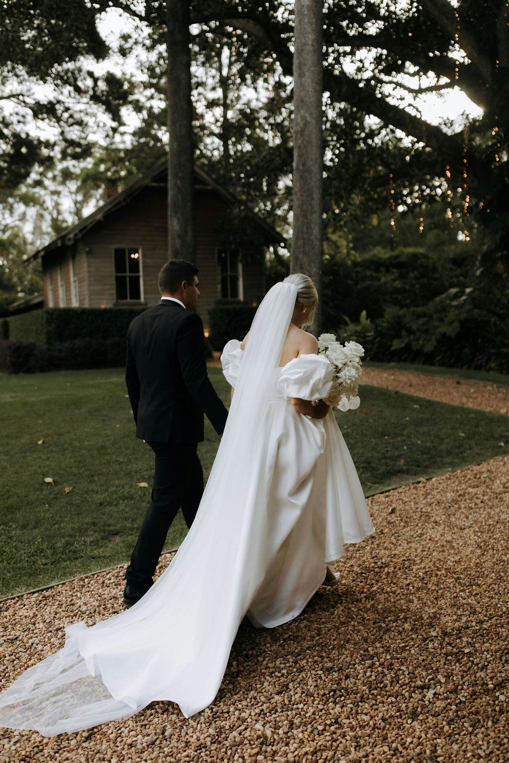 A couple is walking on a gravel path toward a wooden house amidst a lush, green landscape. The bride is wearing a long white gown with a train and holding a bouquet of flowers, while the groom is dressed in a black suit.