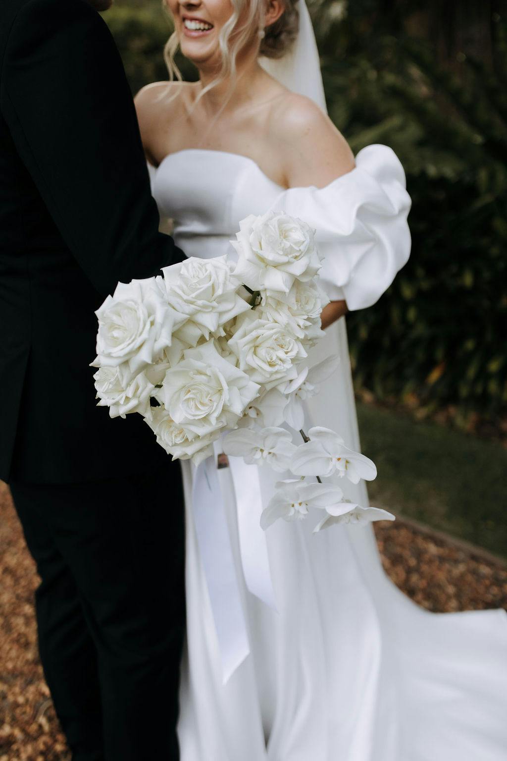 A bride in a white dress holds a bouquet of white roses and flowers while standing next to a groom dressed in black. The background features greenery and a natural setting.