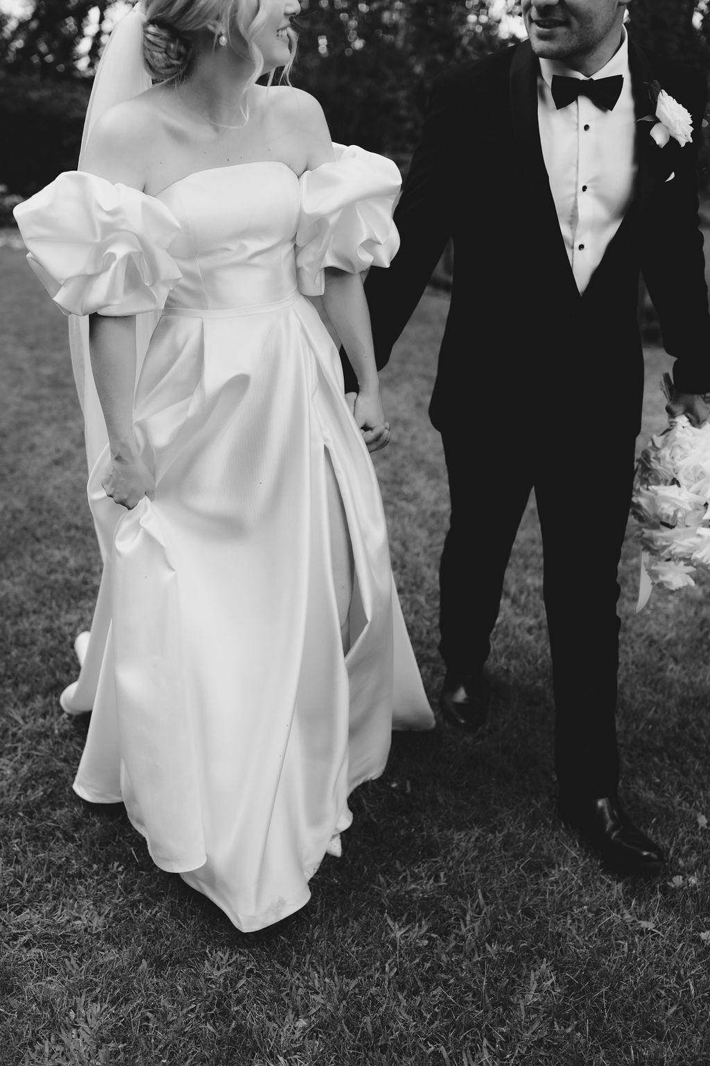 A black-and-white photo of a newlywed couple walking hand in hand on grass. The bride wears an off-shoulder wedding dress with puffy sleeves and holds her gown with one hand. The groom is in a tuxedo with a white shirt and bow tie, holding a bouquet in his other hand.