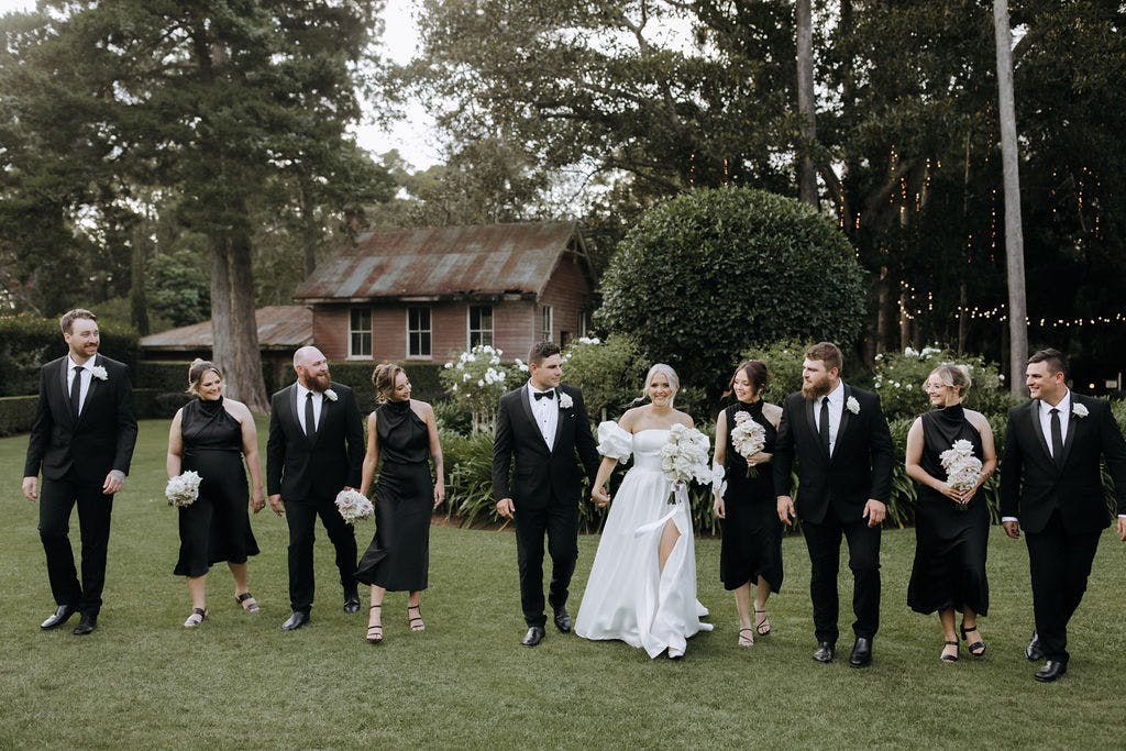 A bride and groom walk with their wedding party on a green lawn. The bride wears a white gown and veil, the groom a black tuxedo. The bridesmaids wear black dresses and carry white bouquets, and the groomsmen wear black suits. Trees and a rustic building are in the background.