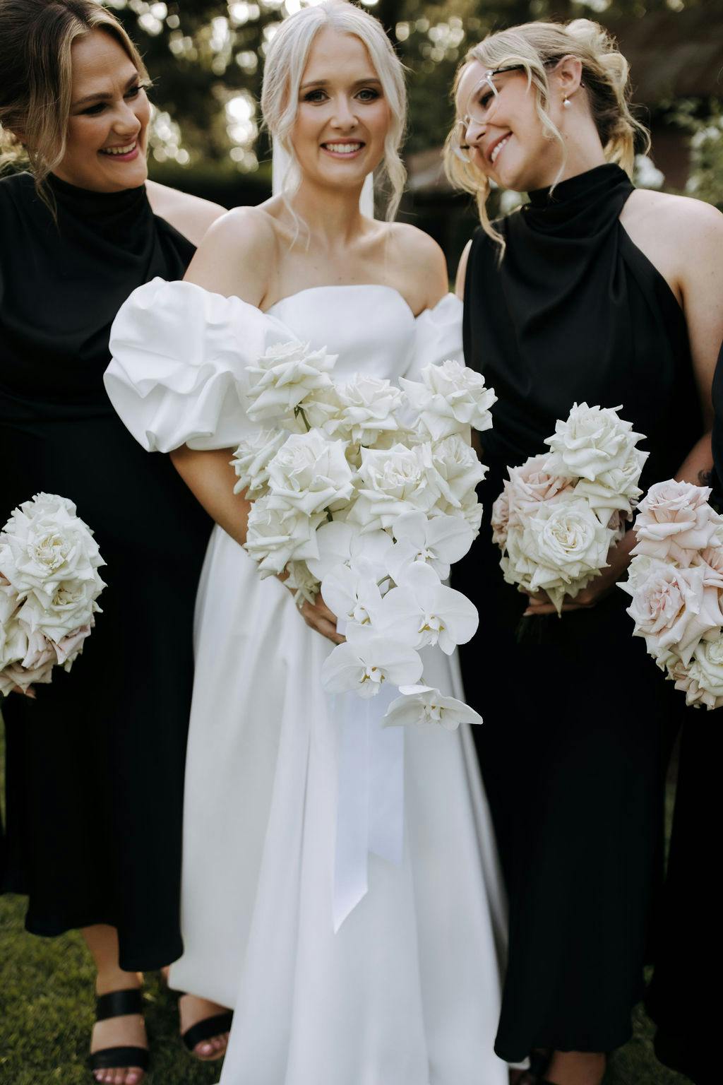 A bride, dressed in a white off-the-shoulder gown with puffed sleeves, holds a cascading bouquet of white flowers. She is flanked by two bridesmaids in black dresses, each holding bouquets of light pink roses. The group is outdoors with greenery in the background.