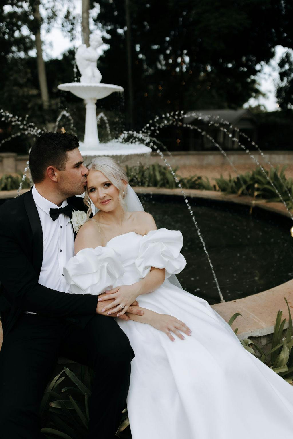 A couple dressed in wedding attire sits by a fountain. The groom, in a black tuxedo, gently kisses the bride, who is wearing an off-shoulder white gown. They are surrounded by greenery, and the fountain water arcs elegantly in the background.