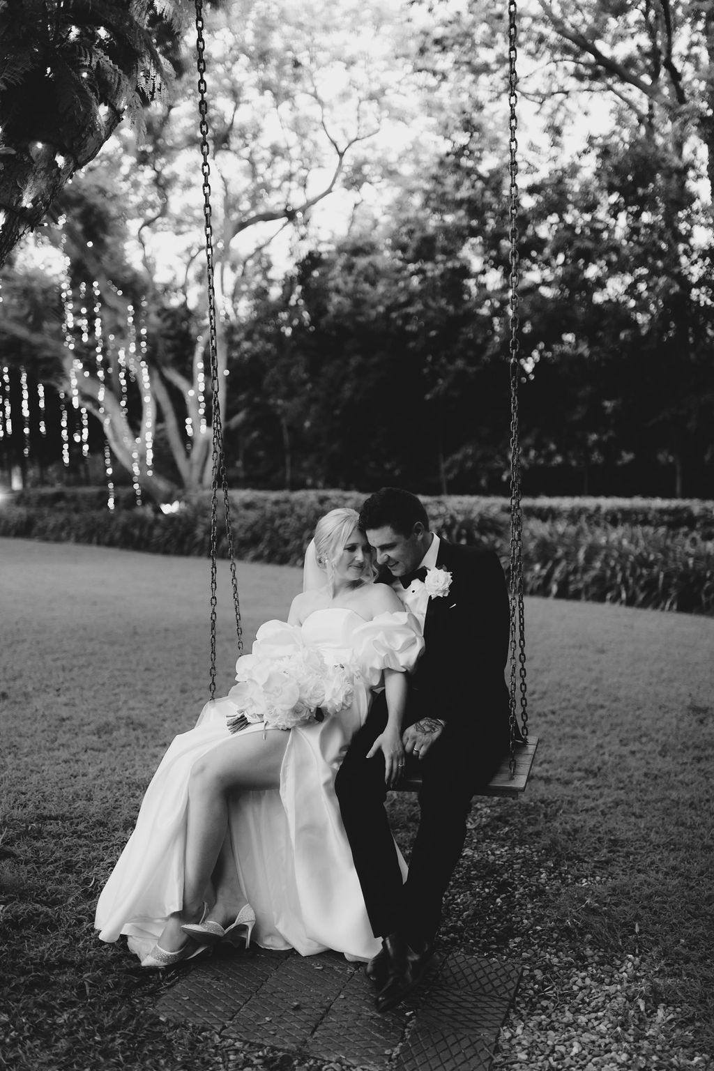 A bride and groom sit closely on a wooden swing in an outdoor setting. The bride, wearing a flowing gown, holds a bouquet and rests against the groom. The groom, dressed in a suit, relaxes next to her. The background features trees and string lights. (Black and white image).