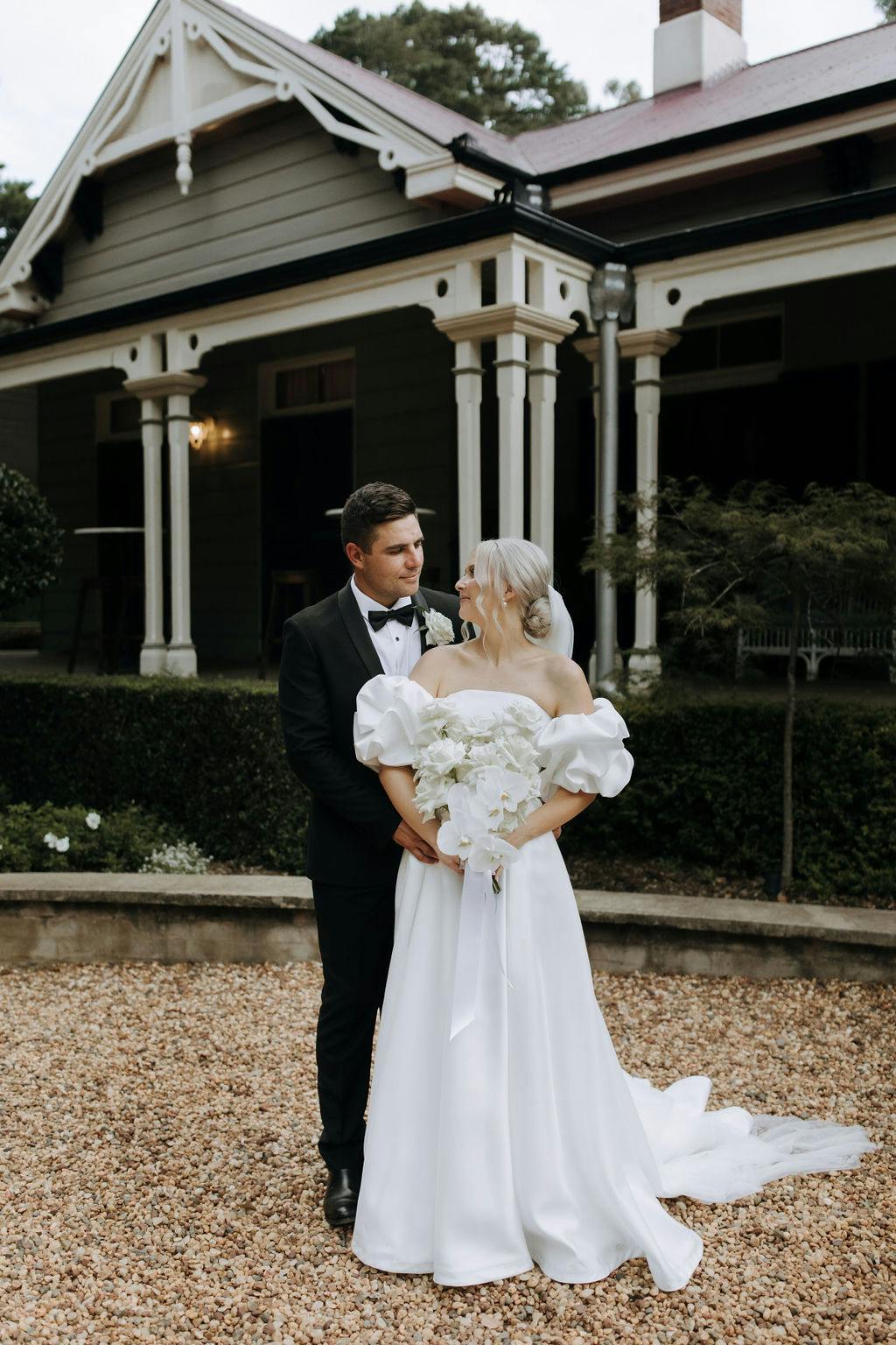 A bride in an off-shoulder white gown holding a bouquet stands next to a groom in a black tuxedo. They are gazing at each other lovingly. They are outside a large, rustic house with a porch and decorative columns, amidst a gravel courtyard and greenery.