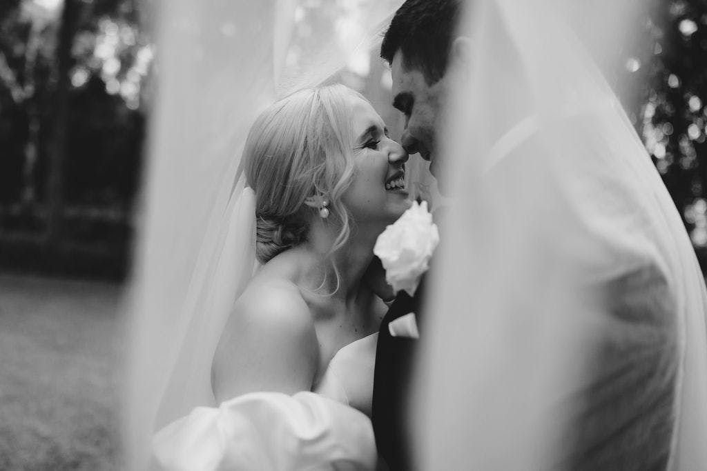 A black-and-white photo of a joyful couple on their wedding day. The bride and groom are standing close, smiling, and gazing into each other's eyes. The bride is wearing a veil, and the groom has a boutonniere. The image captures a tender and intimate moment.