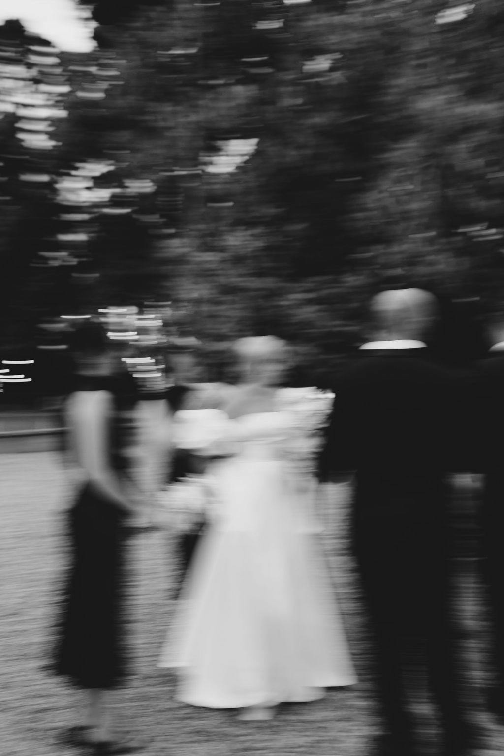 A blurry, black-and-white photo capturing a group of people dressed formally. They appear to be outdoors, possibly in a garden or park, engaged in conversation. The central figure seems to be wearing a white dress, suggesting a wedding scene.