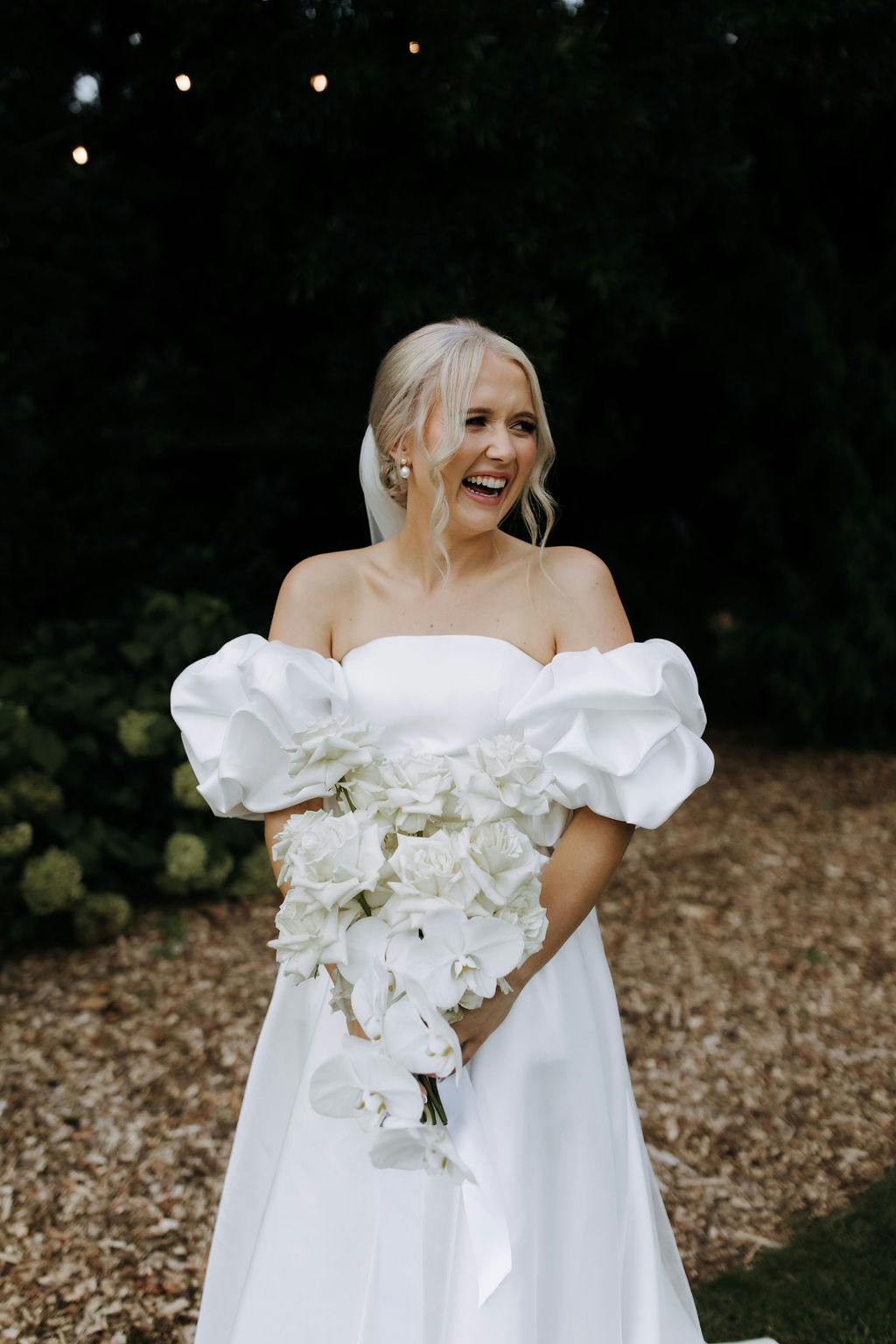 A bride with blonde hair styled in loose waves stands outdoors, smiling brightly. She wears an off-the-shoulder white wedding dress with puffed sleeves and holds a bouquet of white flowers. Behind her, there is greenery and a natural, rustic ground cover of wood chips.