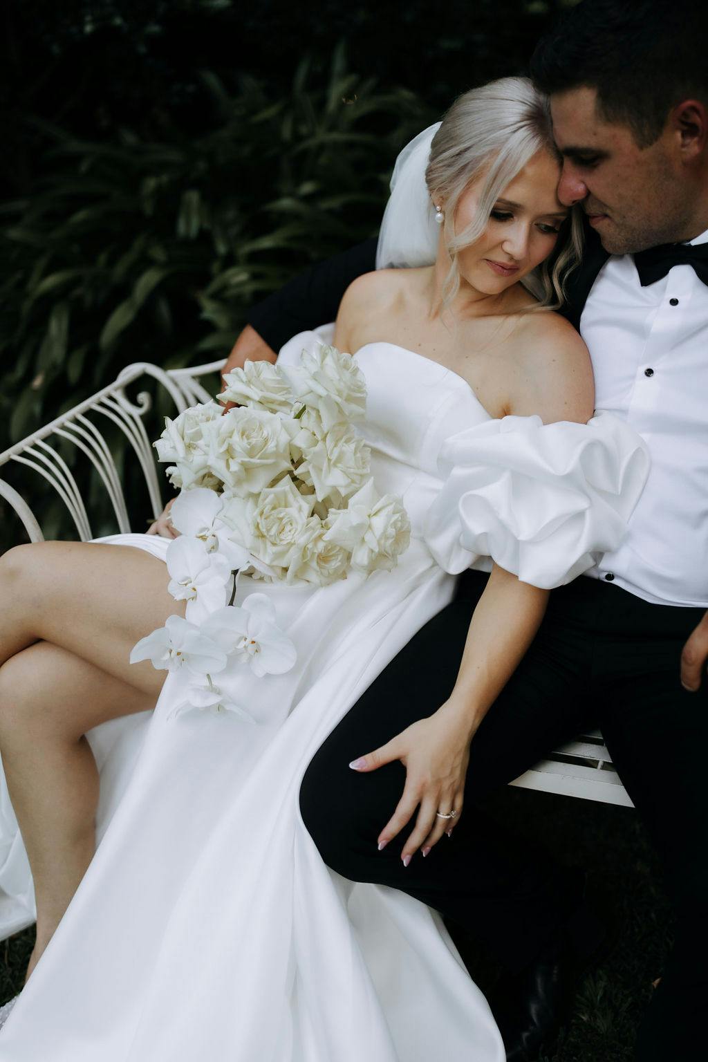 A bride in a white gown with puffy sleeves holds a bouquet of white flowers as she sits close to a groom in a black tuxedo on an outdoor bench. They are nestled together, sharing an intimate and tender moment.