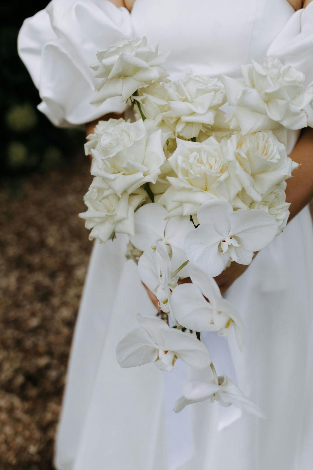 A bride in a white dress holds a bouquet of white roses and white orchids. The flowers are arranged elegantly, creating a classic and timeless look. The background is blurred, focusing attention on the bouquet.