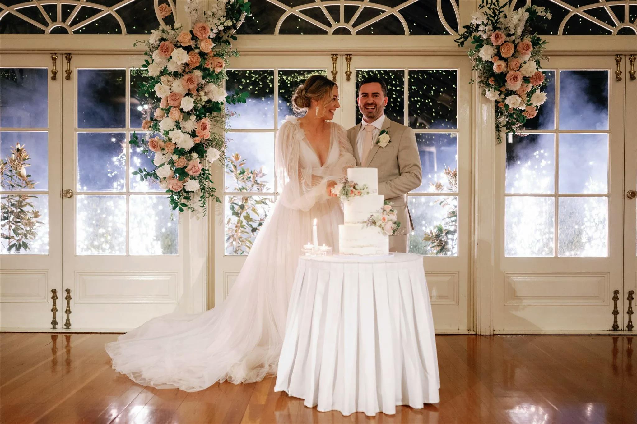 A bride and groom are standing behind a table with a wedding cake in an elegantly decorated room. They are smiling, and the bride is wearing a long white dress while the groom is in a beige suit. The background features large windows with floral arrangements and twinkling lights.
