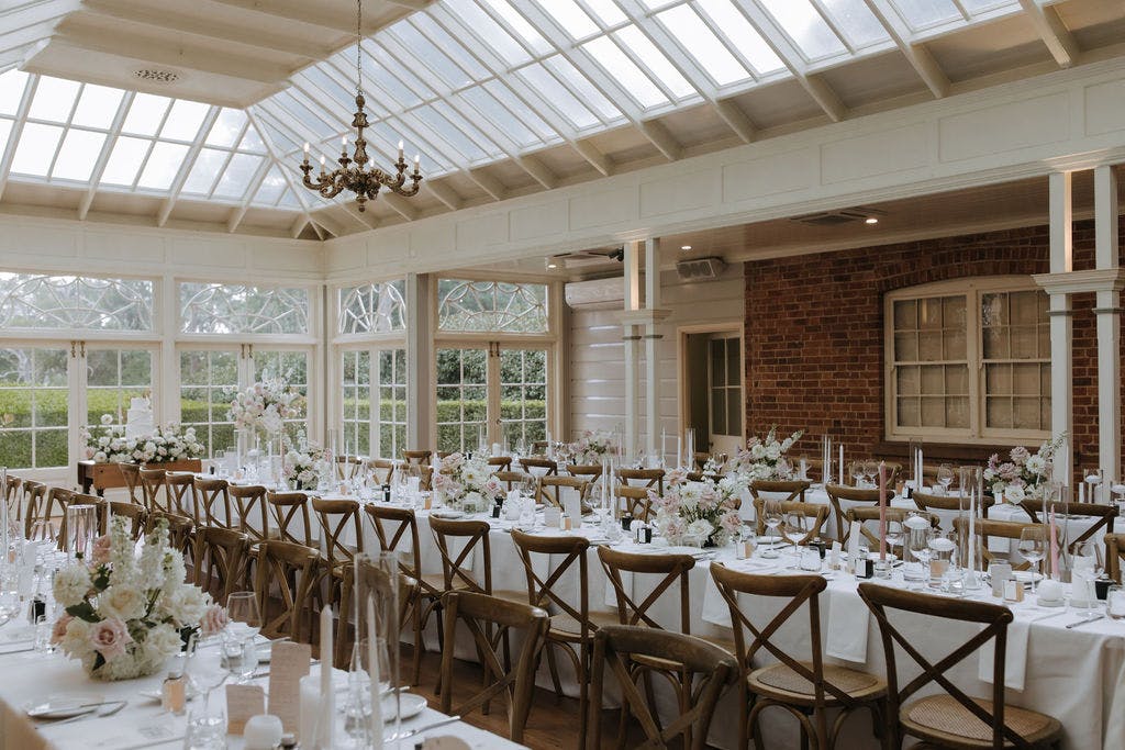 A beautifully decorated wedding reception venue with long banquet tables arranged with white linens, elegant floral centerpieces, and candles. Wooden cross-back chairs surround the tables, under a bright, skylit ceiling with large windows and a chandelier.