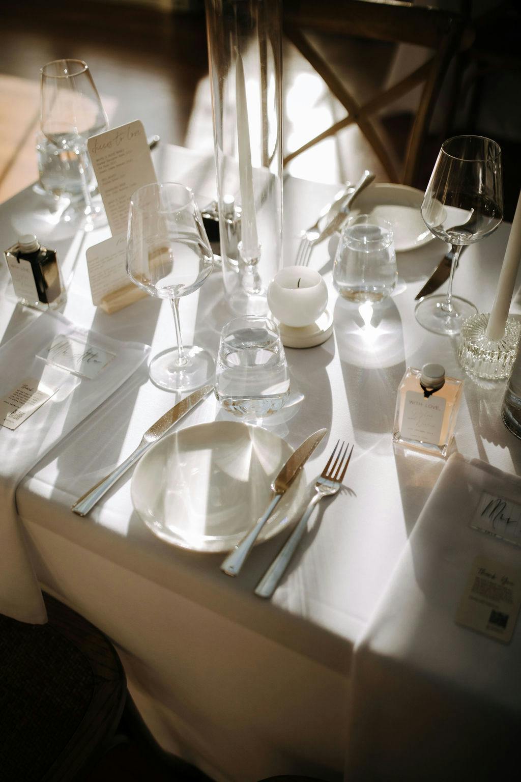 A beautifully set dining table with white linens, glassware, silverware, a candle, and menus. There are empty wine and water glasses, a small bottle of perfume, and a folded napkin on the table, illuminated by soft natural light.